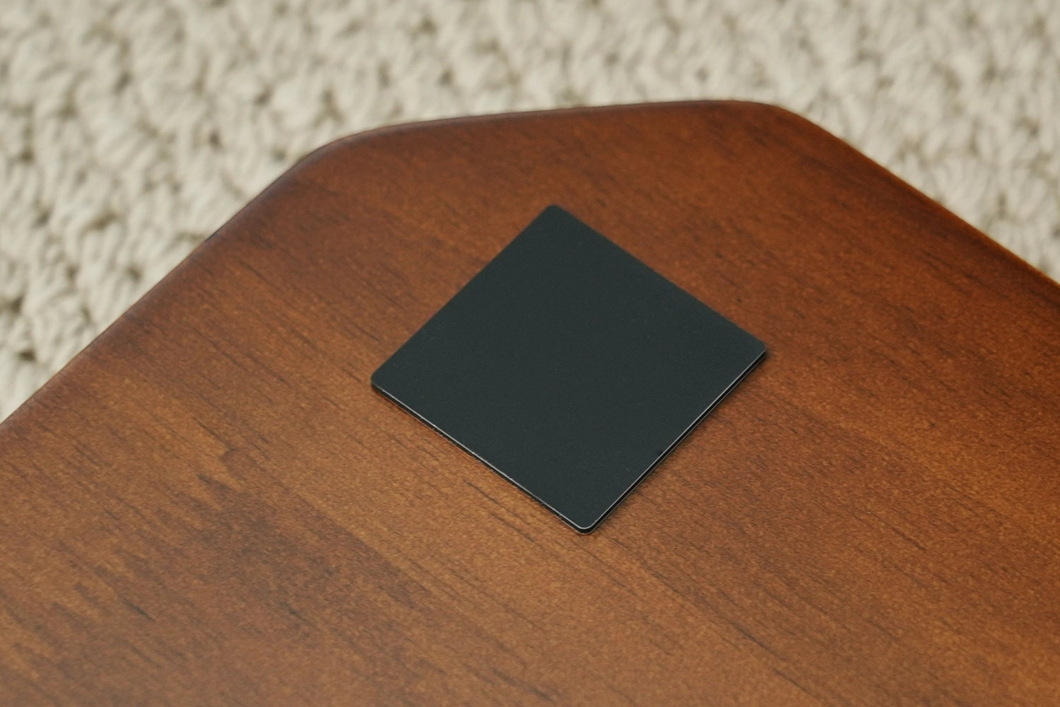 A square black coaster or mat with rounded corners resting on a polished brown wooden surface